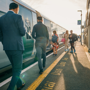 Commuters walking along a sunlit station platform beside a train near Seven Mills