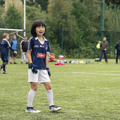 A young girl wearing a mouthguard and GAA sports uniform smiling on a green pitch