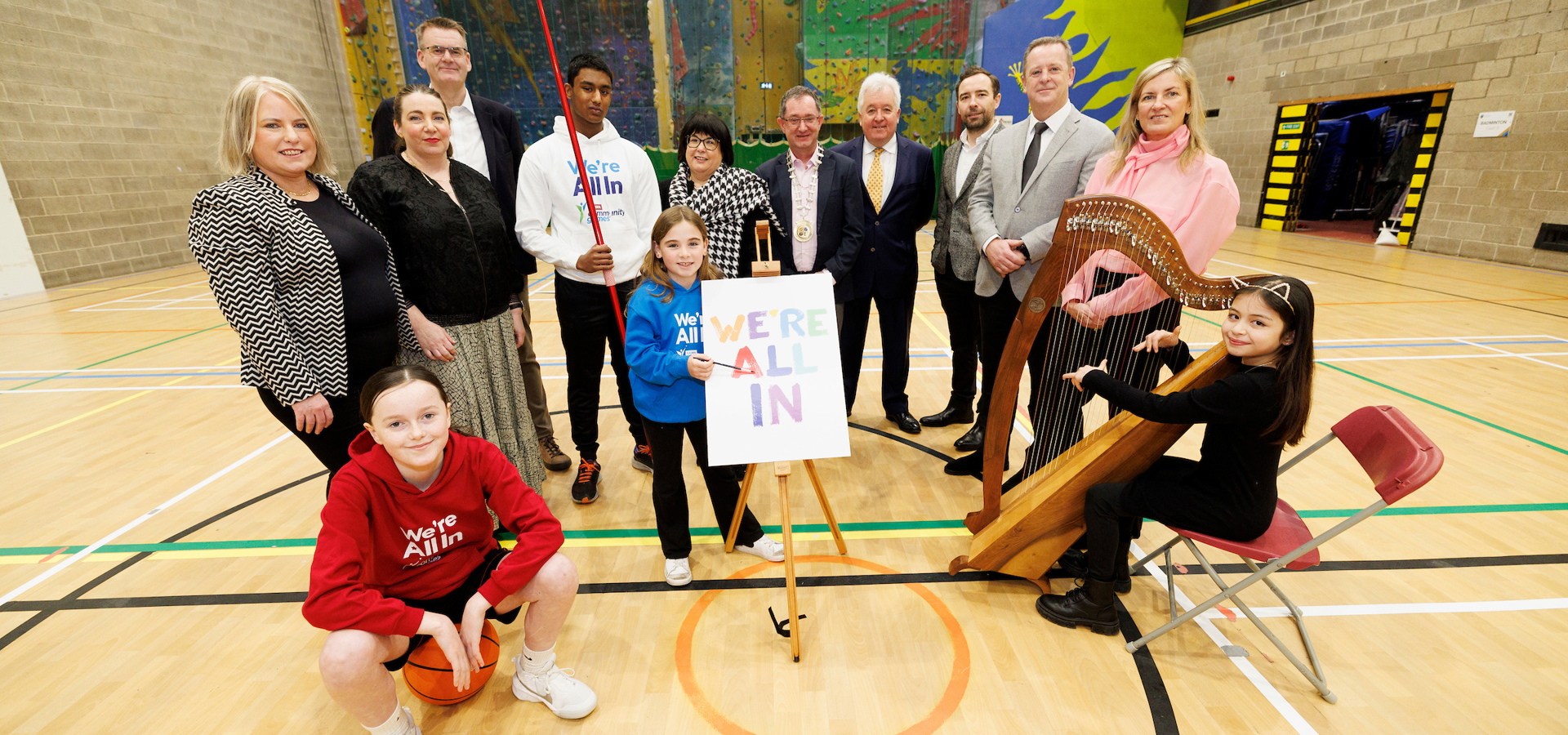 Children with a basketball, harp and javelin beside a 'We're All In' sign at the Community Games launch
